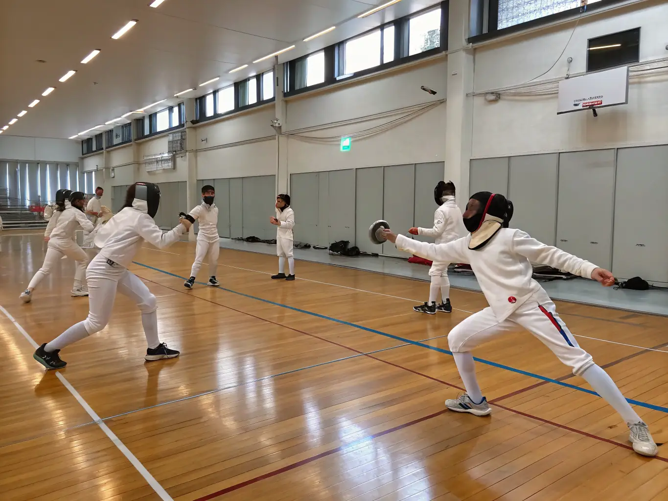 A photograph capturing a group of young fencers participating in a training session, focusing on footwork drills and blade control, set in a modern fencing hall with supportive coaches overseeing the activity.